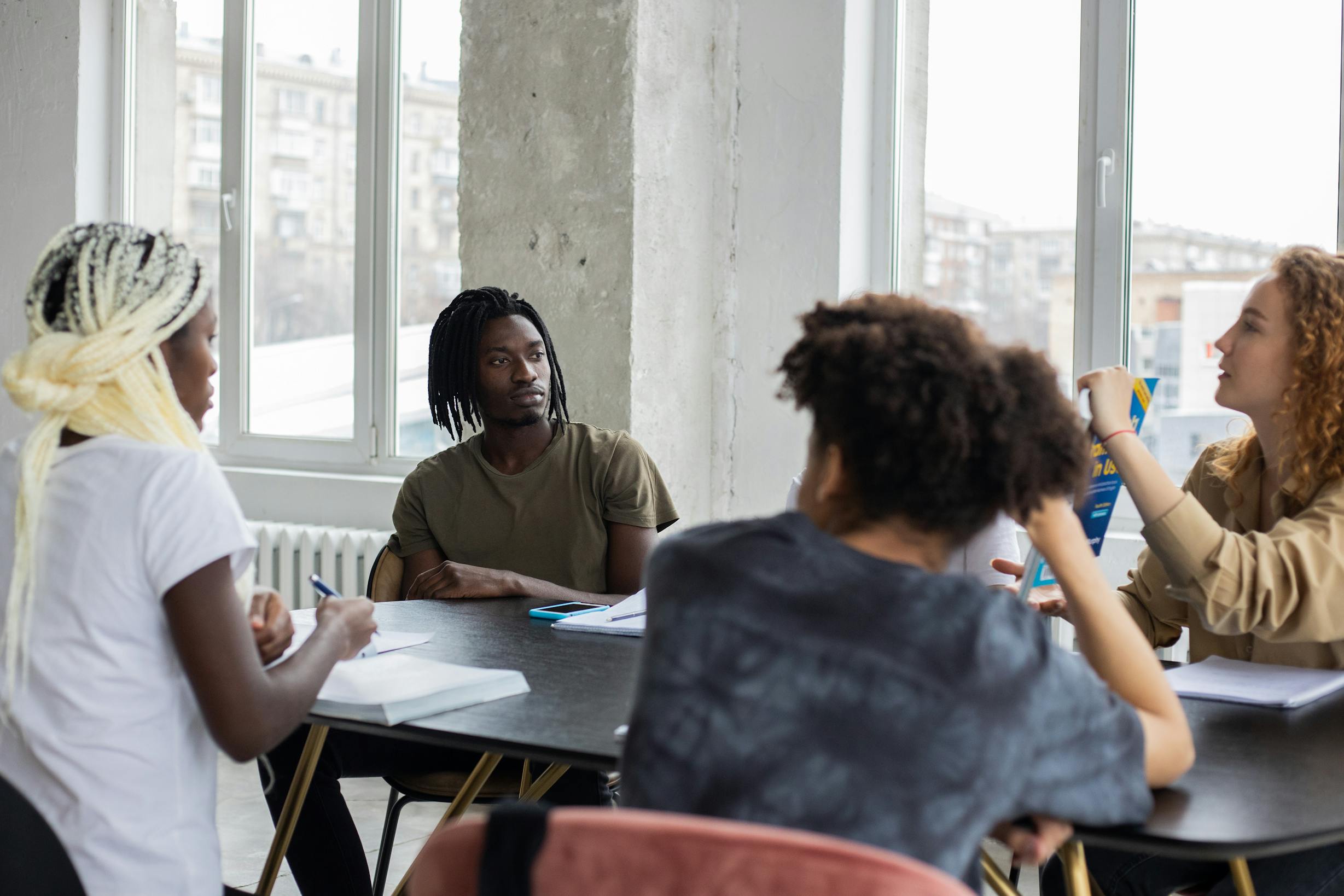 Group of multiethnic students sitting at table with notebooks and book while preparing for exam together in room during lesson